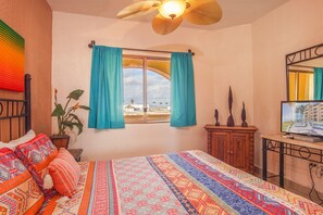 Ein Schlafzimmer mit einem Bett, einem Fenster mit Vorhängen, einem Deckenventilator, einer Pflanze, einem Holzschrank und einem Fernseher.