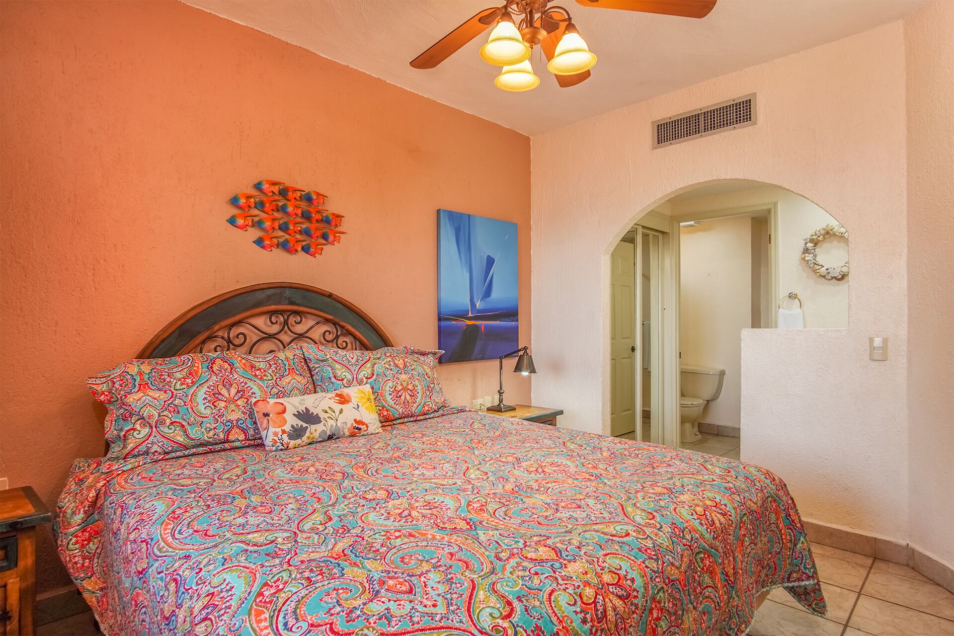 A bedroom with a bed, a nightstand, a ceiling fan, a painting, and an arched doorway leading to a bathroom.