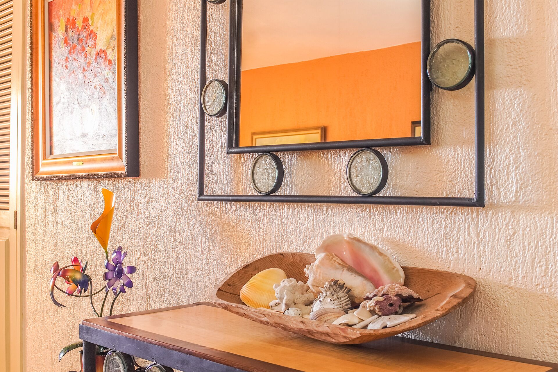 A wooden tray with seashells and a decorative piece on a table, with a mirror and artwork on the wall.