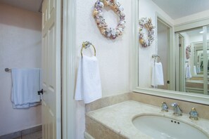 Ein Badezimmer mit einem beigefarbenen Waschbecken, zwei goldenen Wasserhähnen und einem großen Spiegel. Zwei weiße Handtücher hängen an Handtuchhaltern und eine dekorative Muschelkrone schmückt die Wand.