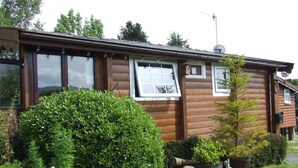 A wooden cabin with multiple windows, a satellite dish, and a well-maintained garden with various plants and flowers.