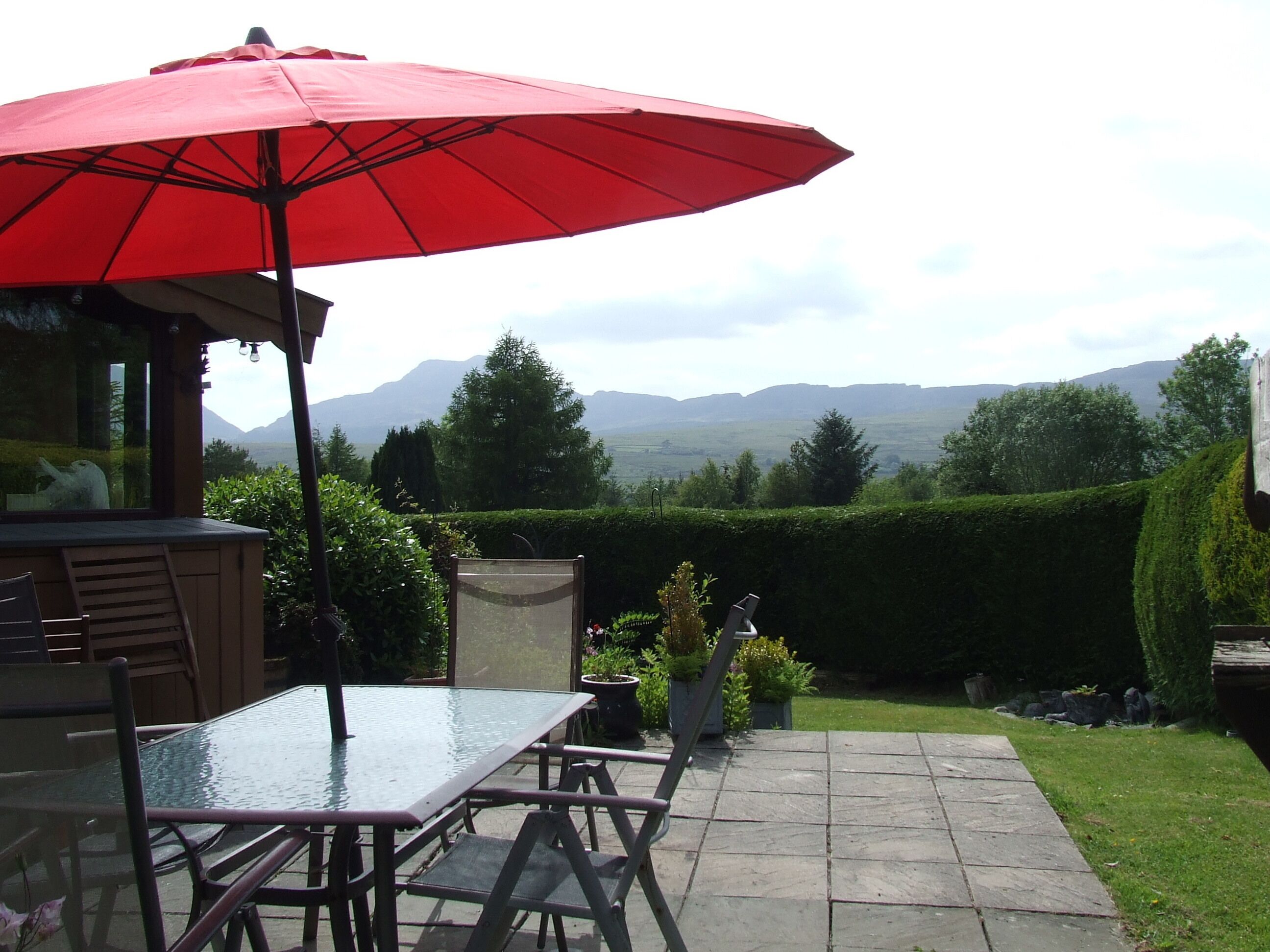 Eine Terrasse mit einem Glastisch und Stühlen, einem roten Sonnenschirm und Blick auf Berge.
