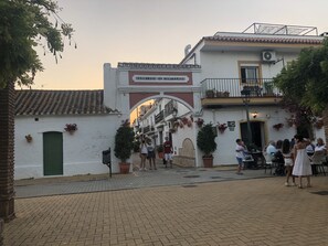Une cour intérieure avec un bâtiment blanc sur lequel on peut lire l’inscription « Barrio de Cancelada ». On y trouve des plantes en pot, des sièges d’extérieur et quelques personnes.