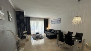 A modern living room with a sofa, a dining table with chairs, a zebra-patterned rug, and a large window with curtains. - Park Beach: Ground floor apartment, garden, nxt to pool, nr beach. Free parking (ESTEPONA  MALAGA)