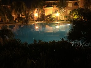 Une vue nocturne d’une piscine entourée de palmiers et d’immeubles illuminés d’une lumière chaleureuse.