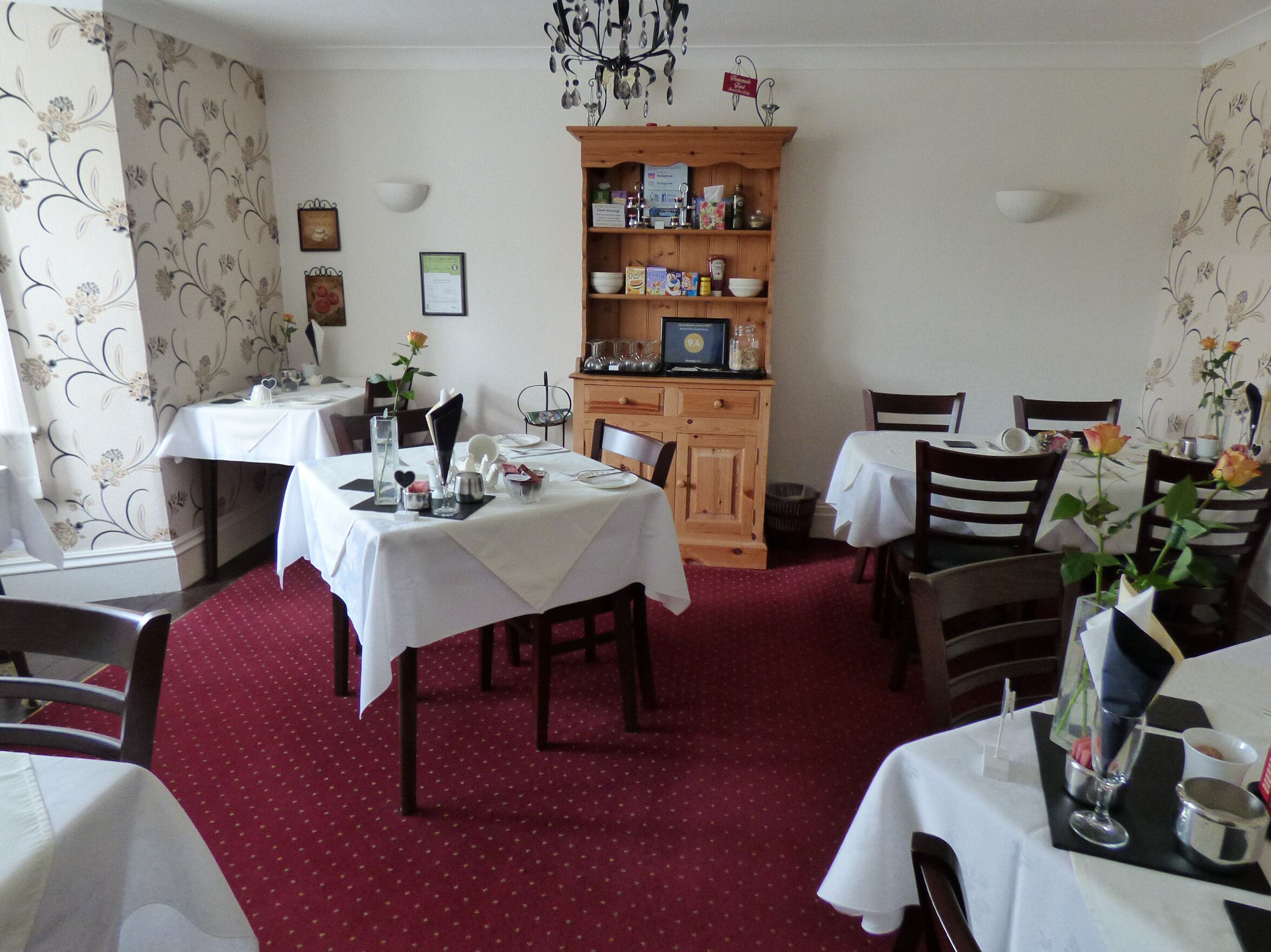 A dining area with tables set for two, a wooden cabinet with various items, and a red carpeted floor.