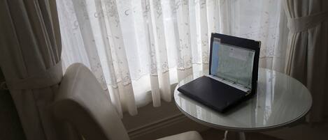 Una computadora portátil sobre una mesa de vidrio cerca de una ventana con cortinas translúcidas.
