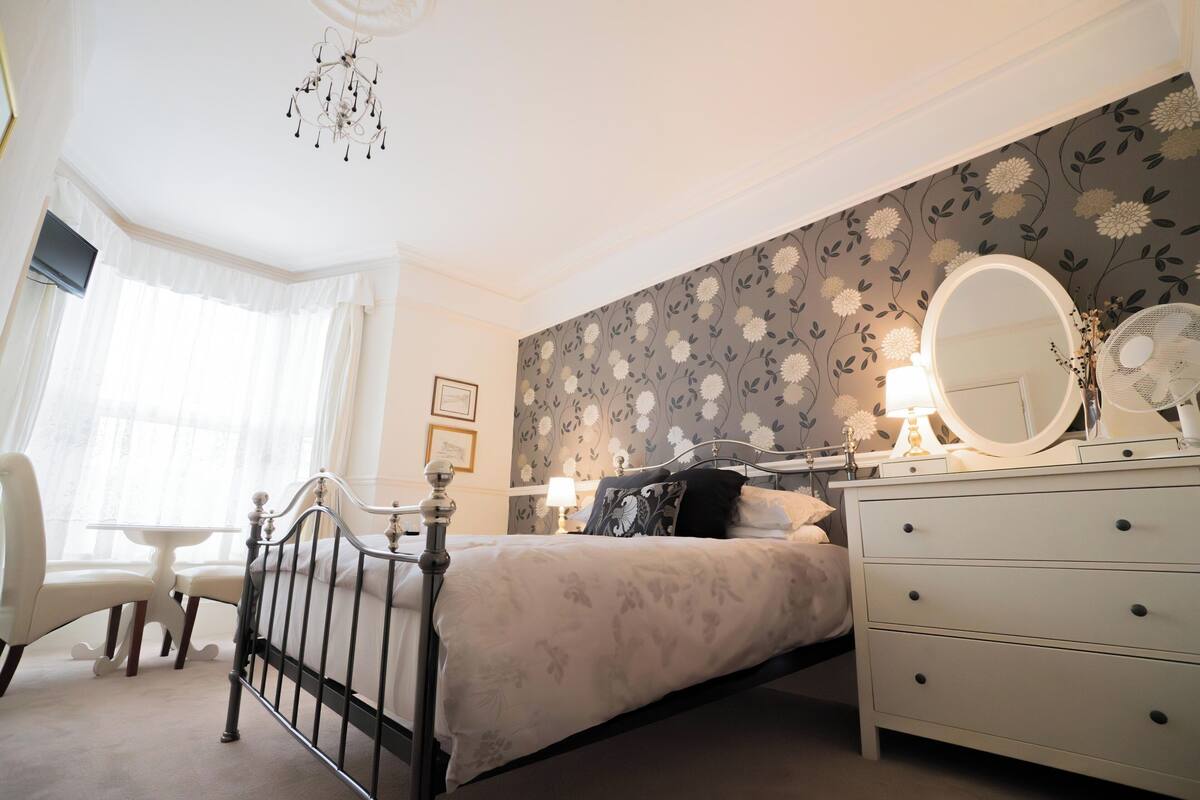 A bedroom with a bed, bedside table, dresser, and a large mirror.