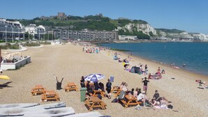 A beach with wooden benches and umbrellas, a clear sky, and buildings in the background. - Maison Dieu Guest House (Dover)