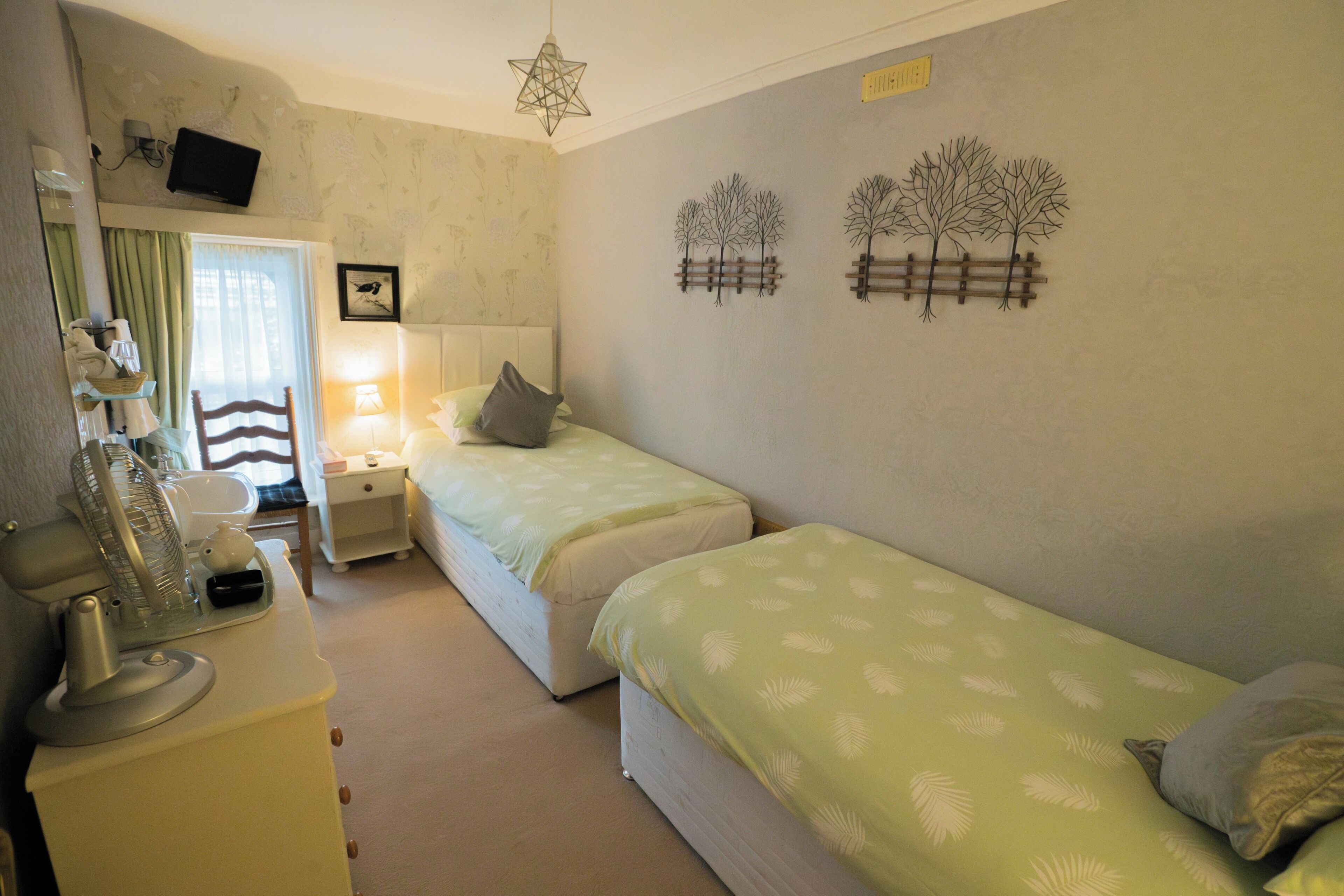 A bedroom with two single beds, a bedside table with a lamp, a dresser, a mirror, and a television mounted on the wall.