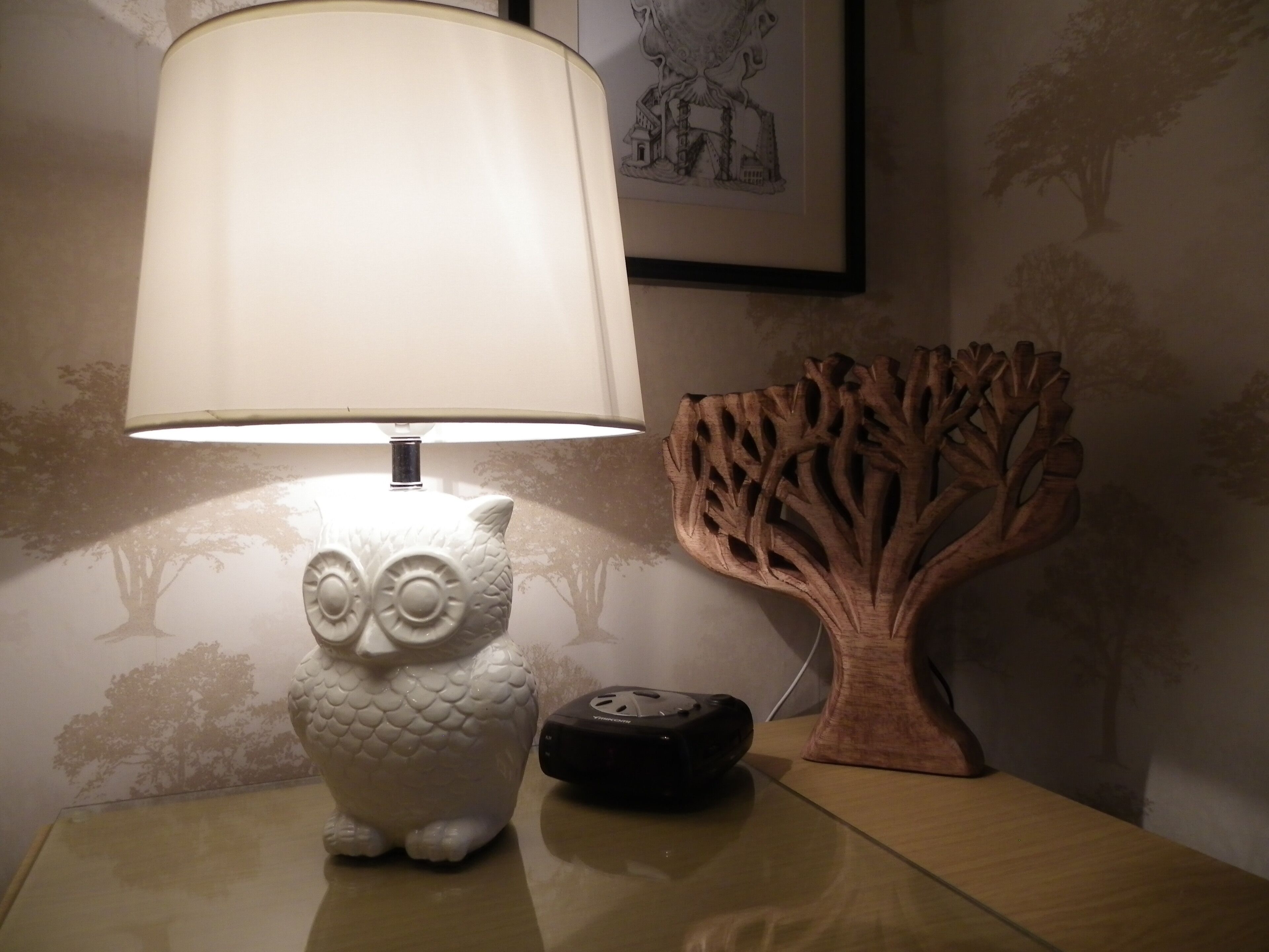 Una lámpara de cerámica blanca con forma de búho sobre una mesita de noche, con una escultura con forma de árbol a su lado.