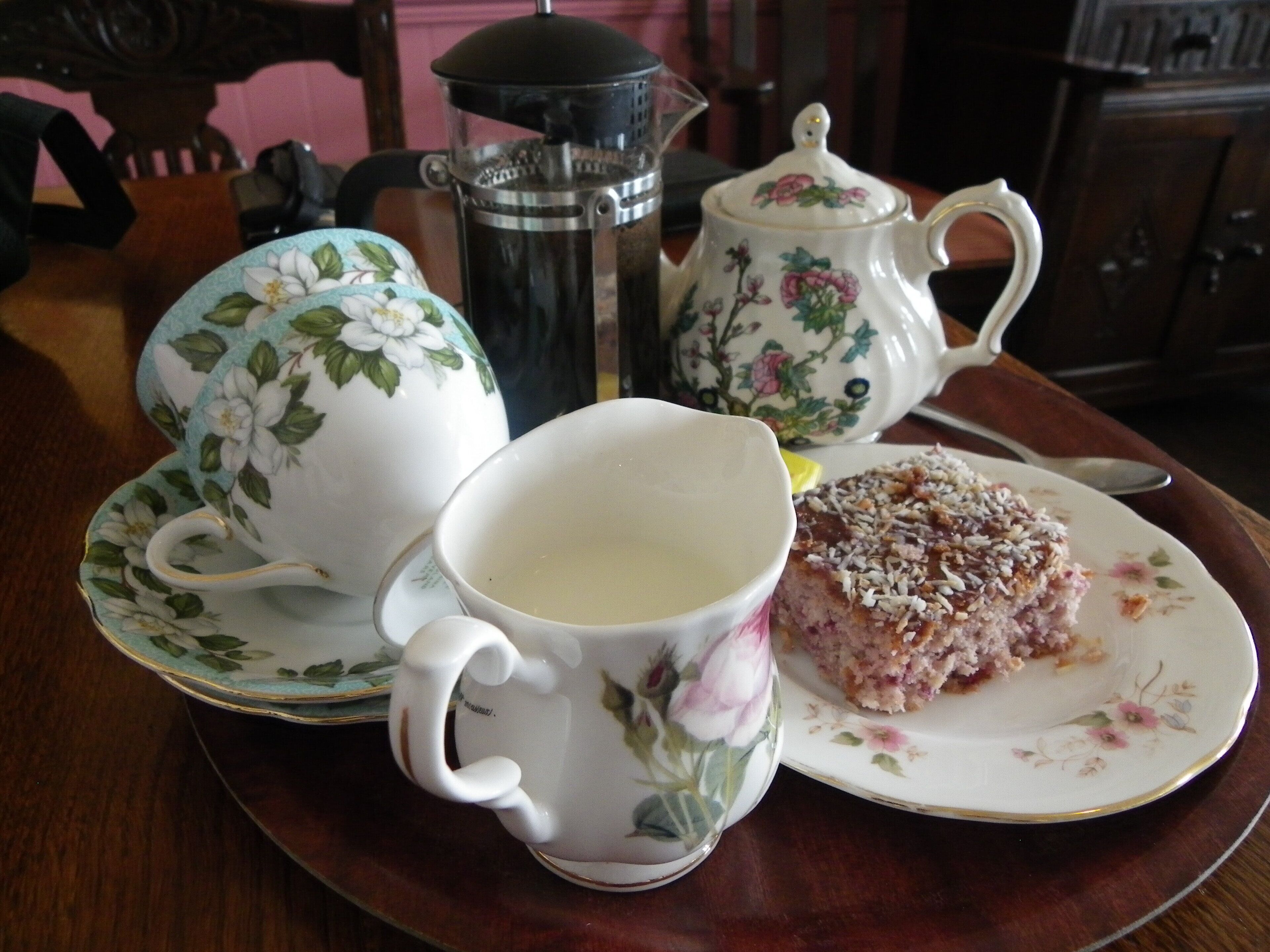 A table with a floral teapot, a coffee pot, a cup of tea, and a slice of cake on a plate.