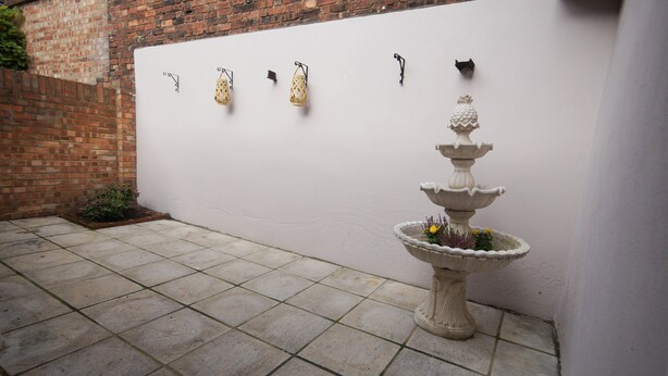 Un patio con un muro blanco, un muro de ladrillo, piso de baldosas y una fuente decorativa con flores.