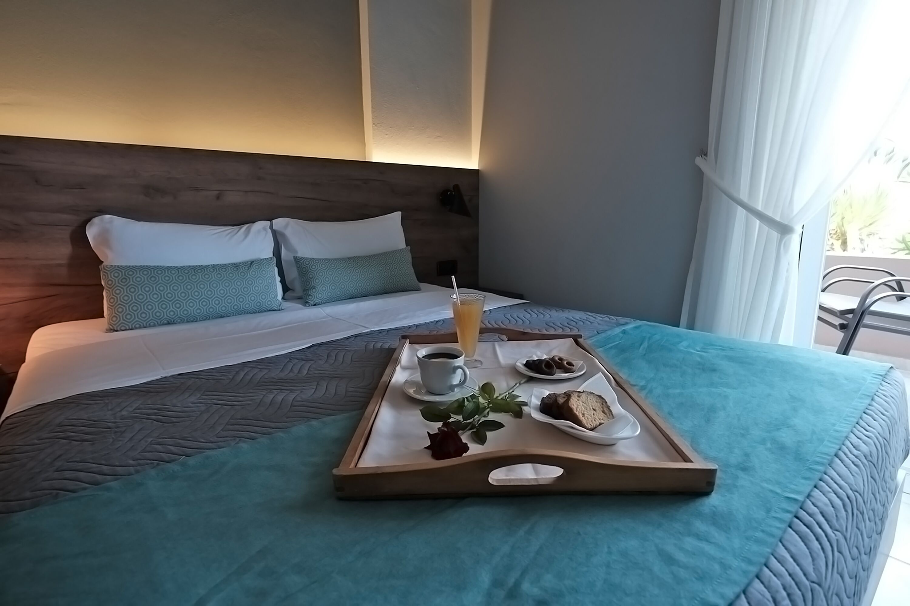 A neatly made bed with a tray of breakfast items including coffee, juice, and pastries, set against a modern room with a wooden headboard and a view of the outdoors.