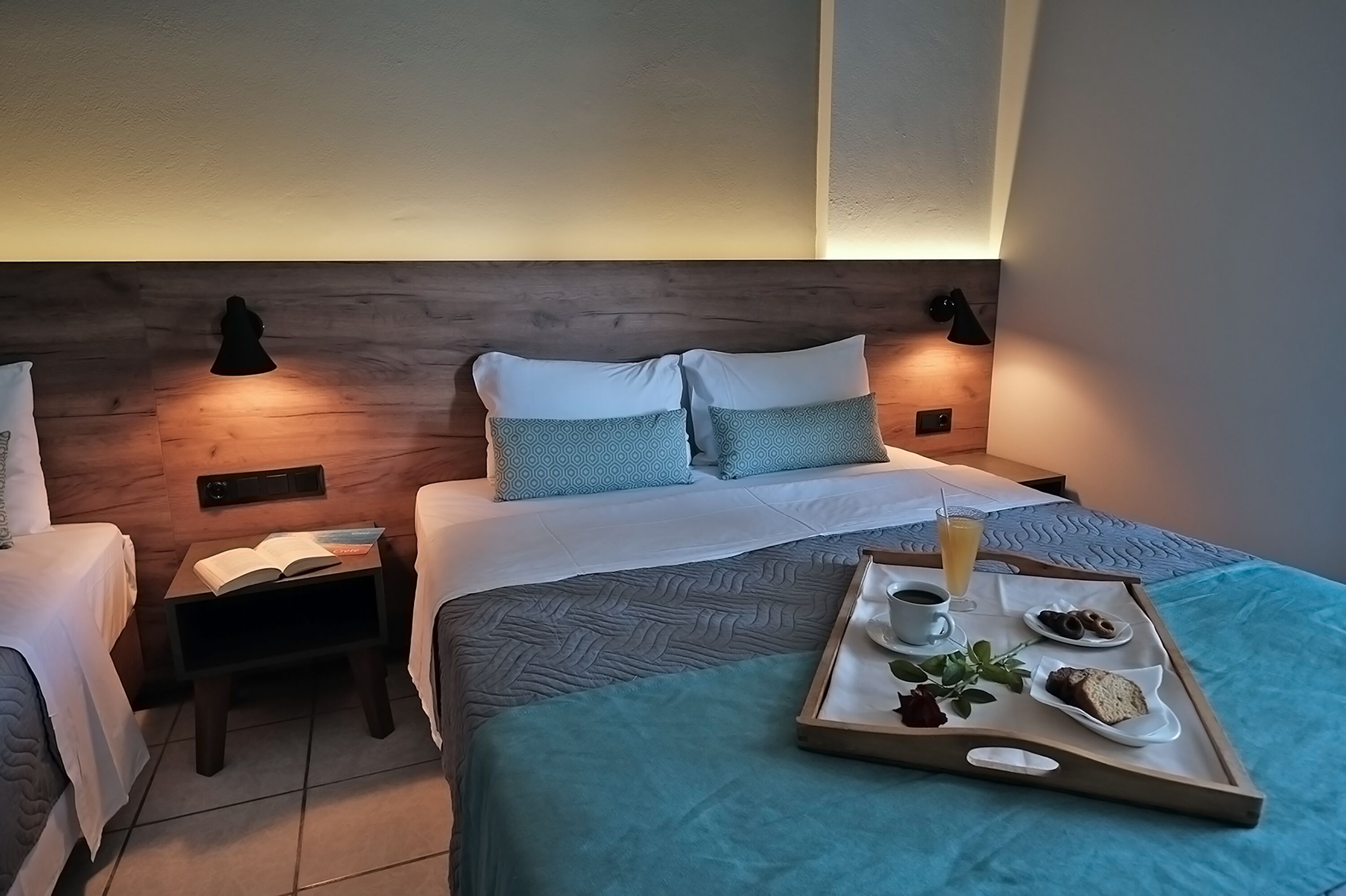 A hotel room with a wooden headboard, two beds with matching pillows, bedside tables with lamps, and a tray with breakfast items.