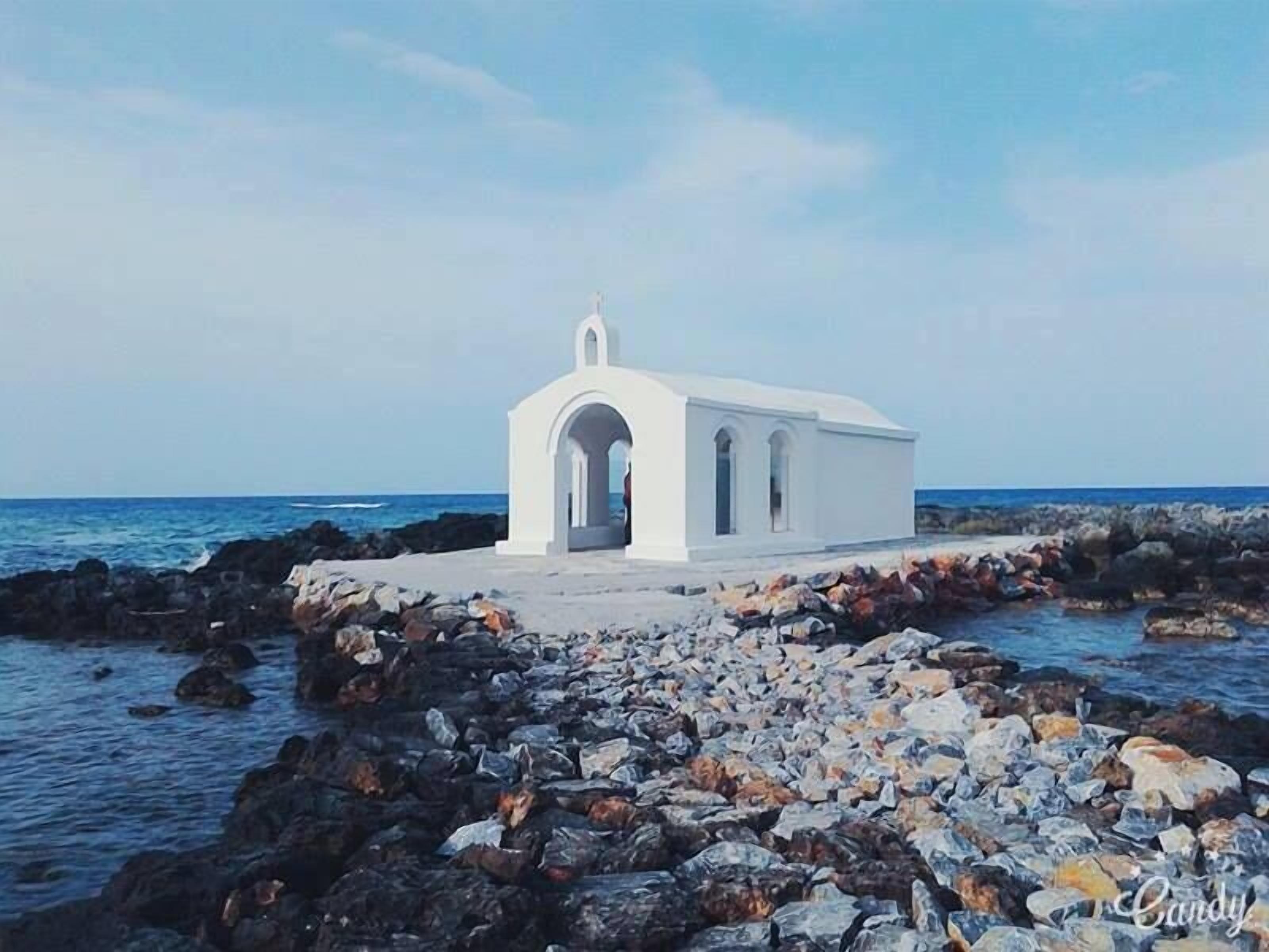 A white building with a small entrance and a bell tower, situated on rocky terrain near the sea.
