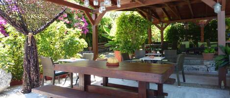 Une terrasse extérieure avec des tables et des bancs en bois, entourée de plantes en pot et agrémentée d’une pergola ornée de lanternes suspendues.