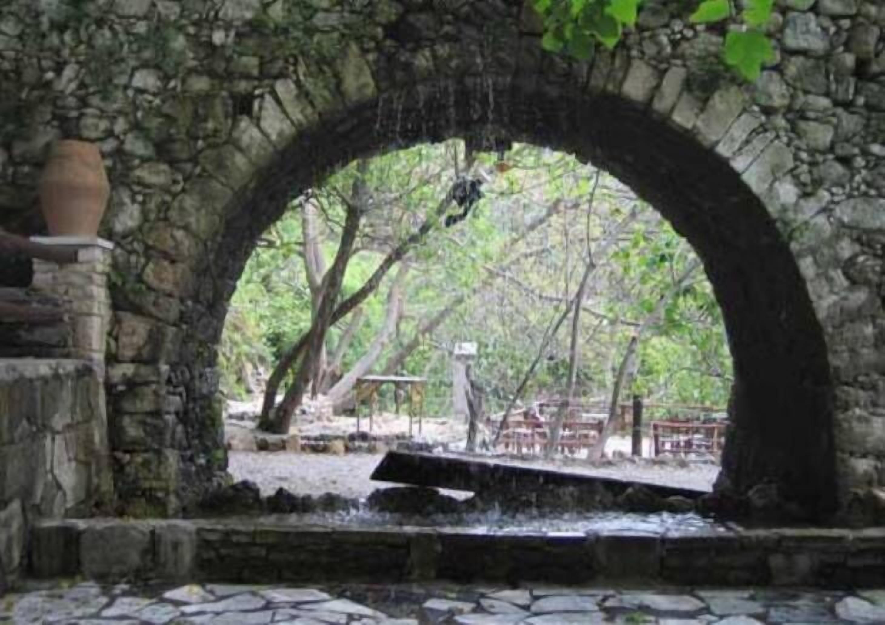 Une structure en pierre en forme d’arche, traversée par un petit ruisseau, entourée d’arbres et agrémentée d’un banc en bois.