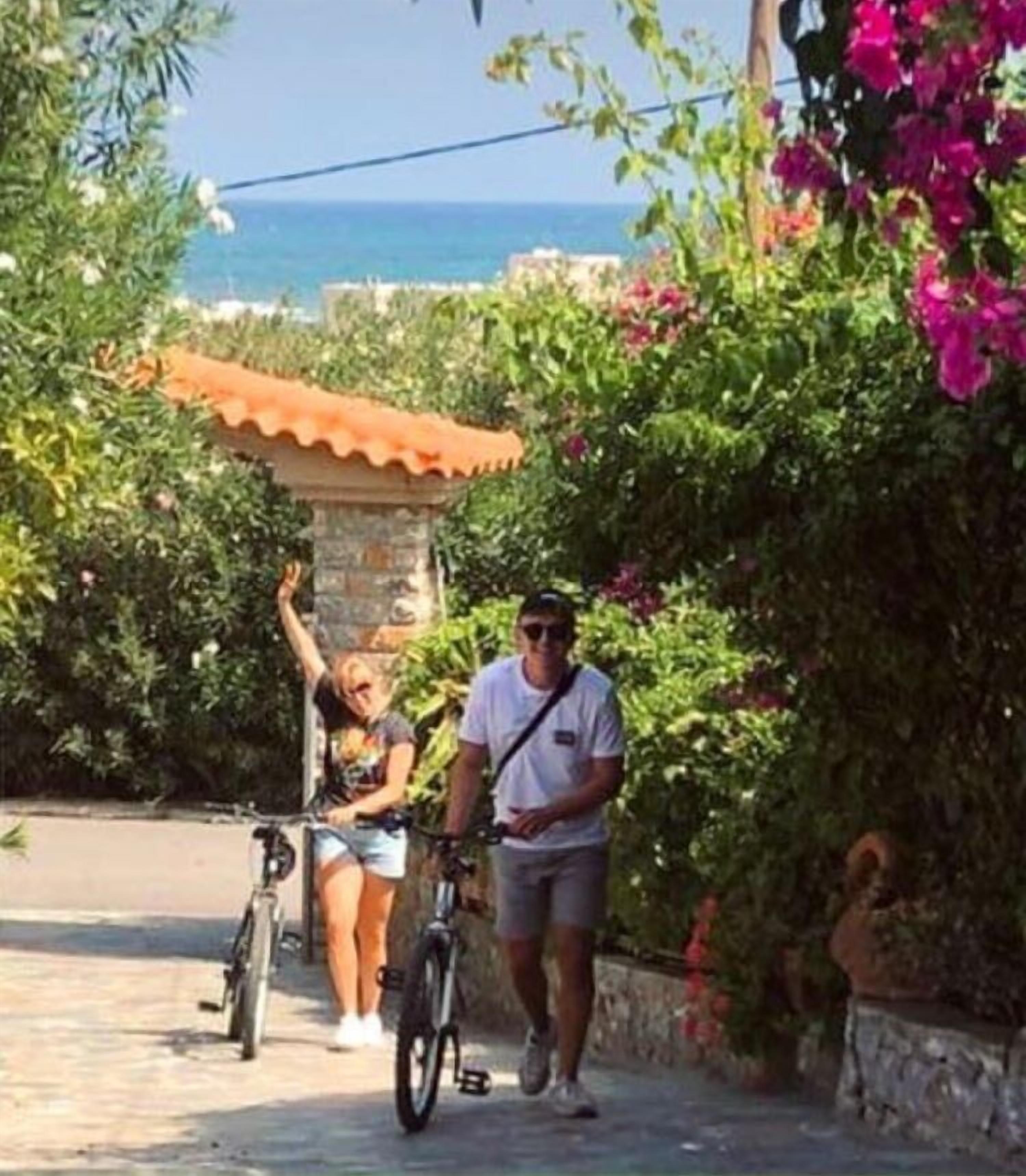Un hombre y una mujer están de pie cerca de una bicicleta, con un muro de piedra y follaje al fondo.