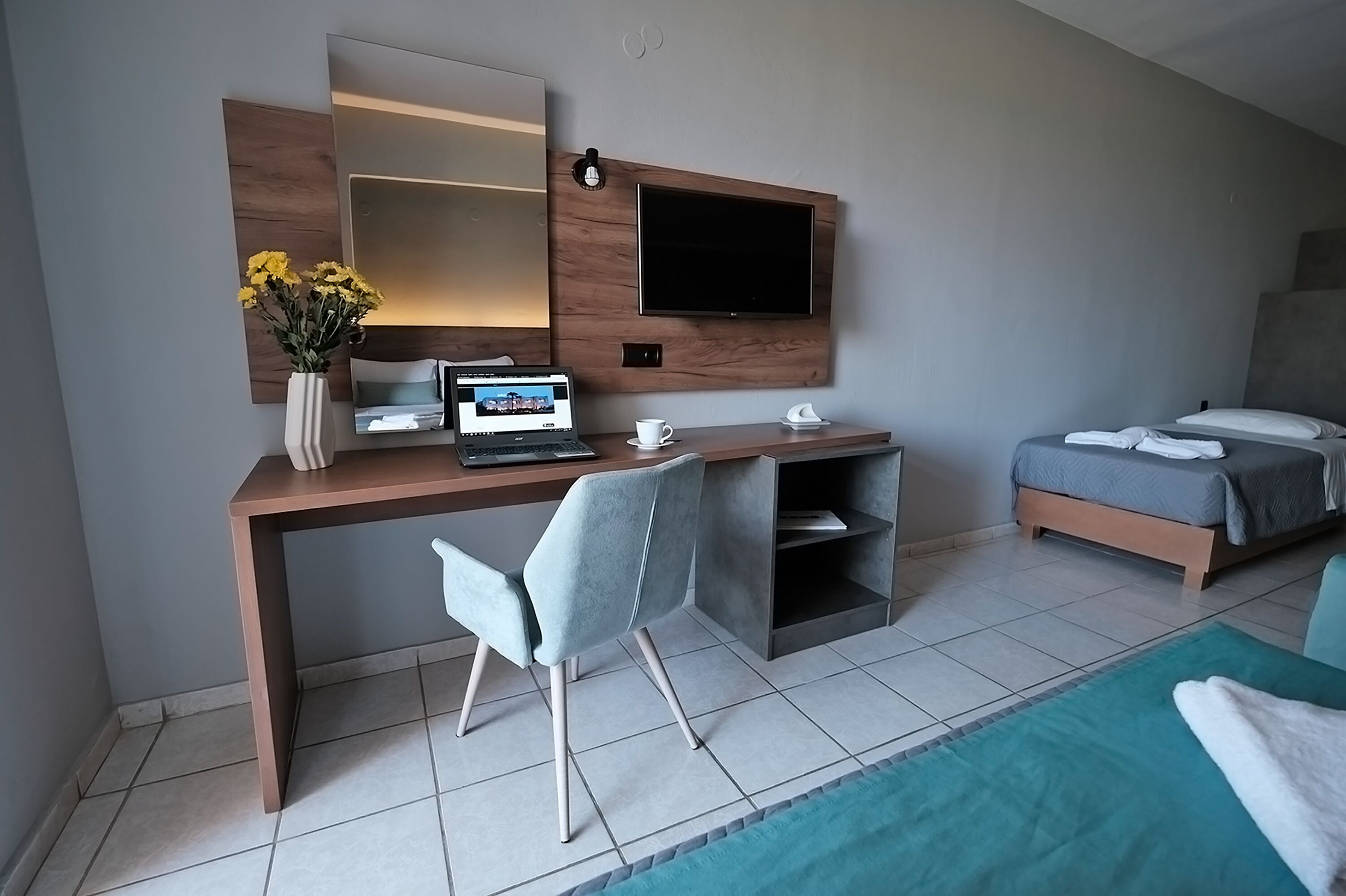 Moderni hotellihuone, jossa on puinen työpöytä, kannettava tietokone, tuoli, harmaalla peitolla varustettu sänky ja seinälle asennettu televisio.