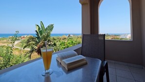 A balcony with a table, a drink, a book, and a view of the sea and greenery. - Poledas Apartments (Apokoronas)