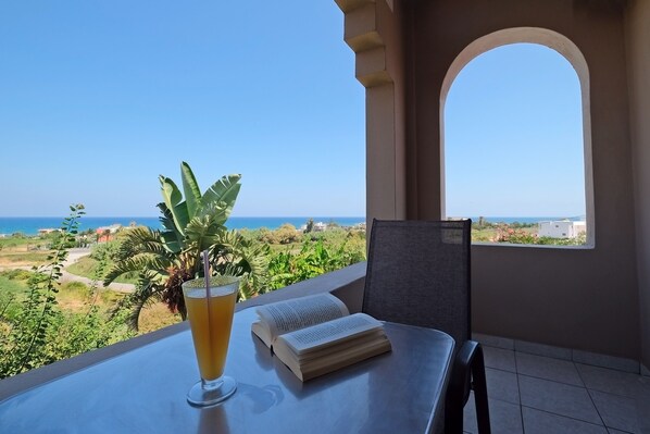 A balcony with a table, a drink, a book, and a view of the sea and greenery. - Poledas Apartments (Apokoronas)