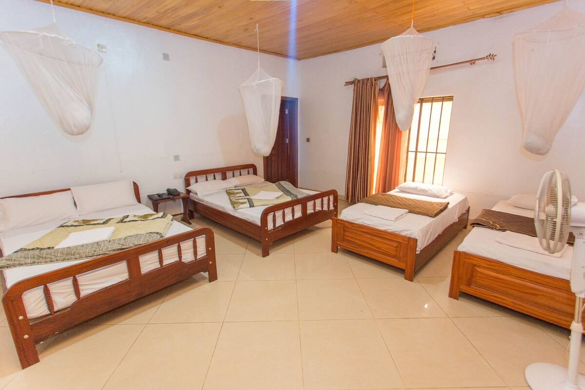 A room with four wooden beds, a standing fan, and mosquito nets on the ceiling.