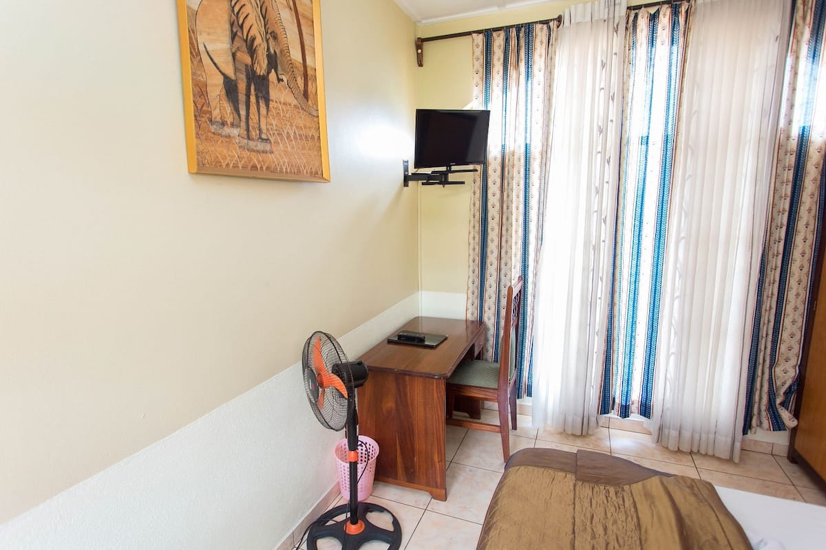A room with a wooden desk, a chair, a bed, a standing fan, a TV mounted on the wall, and a window with curtains.
