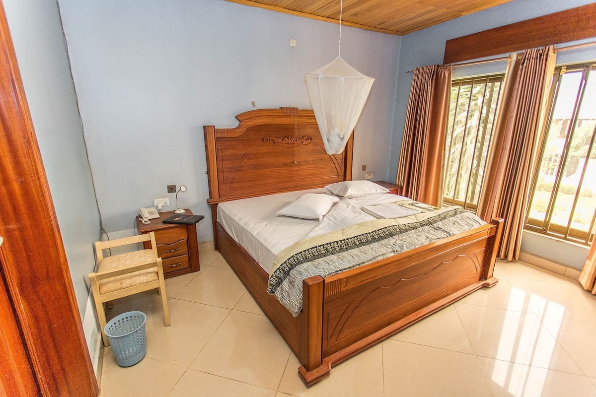 A wooden bed with a mosquito net, a bedside table with a lamp, and a window with curtains.
