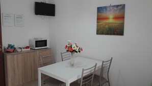 A small dining area with a white table and chairs, a microwave on a wooden cabinet, a vase with flowers, and a painting of a sunset on the wall. - Anita's House in the Heart of Segesta CIN IT081003C2BTVOXQJS  (Calatafimi)