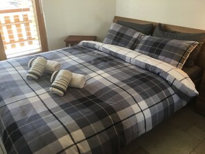 Ein Bett mit karierter Bettdecke, zwei aufgerollte Handtücher und ein Nachttisch daneben.