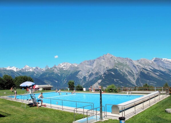 A swimming pool with a mountain backdrop, surrounded by a grassy area and a fenced pool deck.