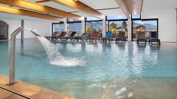 An indoor swimming pool with a water slide, lounge chairs, and a view of mountains through large windows.