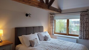 Un dormitorio con una cama grande, una mesita de noche con lámpara, una viga de techo de madera y una ventana con cortinas.