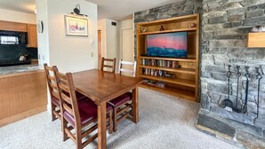 A dining area with a wooden table and chairs, a stone wall with a mounted TV, and a fireplace with metal hooks. - JHRL - Sleeping Indian West #10 (Teton Village)
