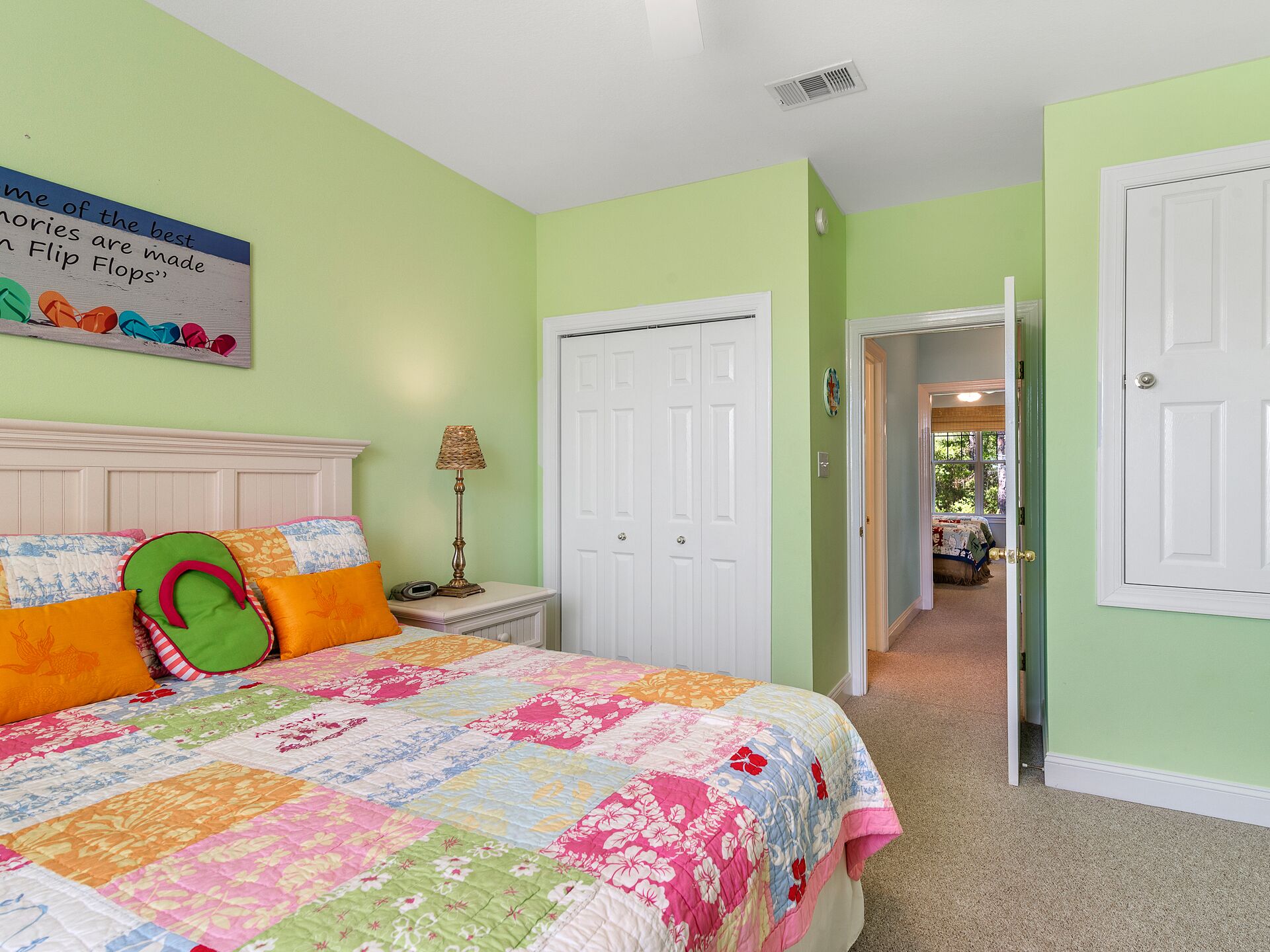 A bedroom with a colorful quilt, a bed with pillows, a nightstand with a lamp, and a door leading to another room.