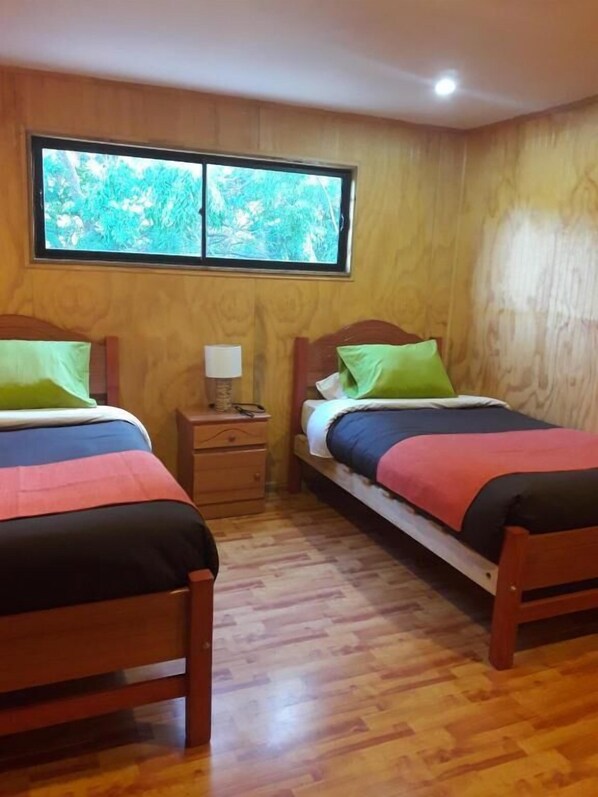 A room with two wooden beds, a nightstand, and a window with a view of greenery. - La Casa del Kori - Hostal - Hostel (Hanga Roa)