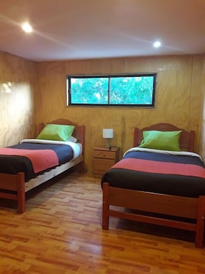 Ein Zimmer mit zwei hölzernen Betten, einem kleinen Nachttisch und einem Fenster mit Blick auf Bäume.