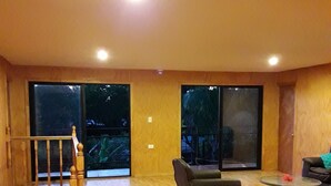 A spacious living area with a wooden floor, a black and white striped rug, a sofa, a coffee table, and a staircase leading to a balcony. - La Casa del Kori - Hostal - Hostel (Hanga Roa)