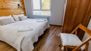 Et soveværelse med sengegavl i træ, en stor seng med hvidt sengetøj, en træstol med pude og et vindue med udsigt til træer.