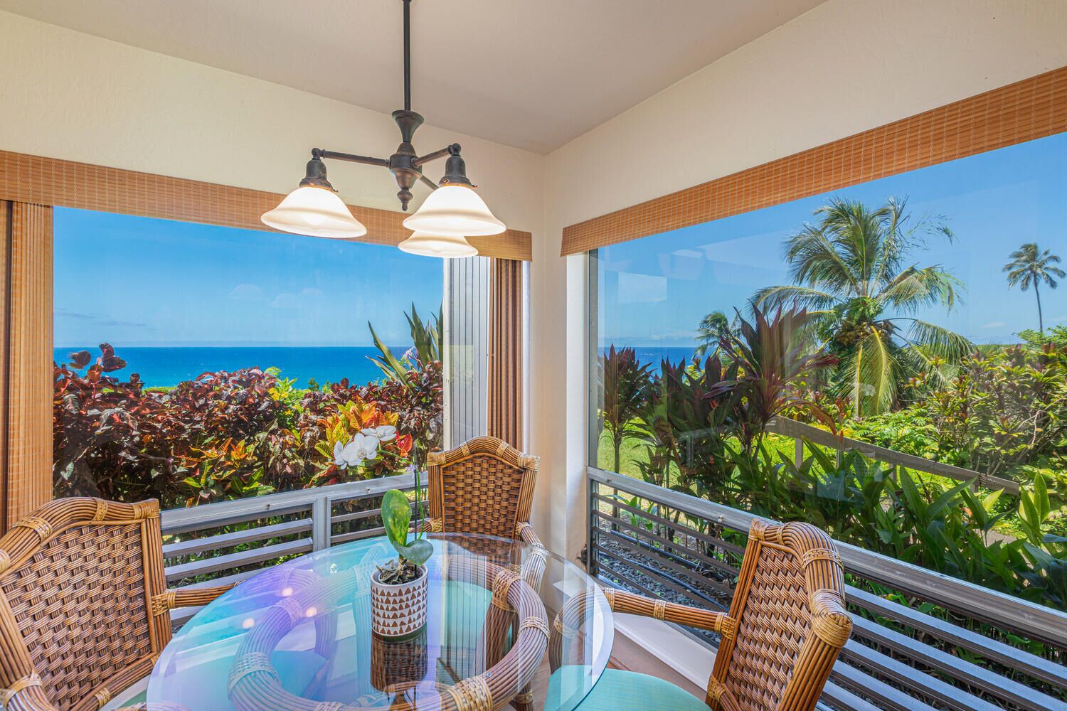 A round glass table with wicker chairs, a potted plant, and a view of the ocean and tropical vegetation through large windows.