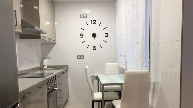A modern kitchen with a built-in clock, stainless steel appliances, and a dining area with white chairs and a table.
