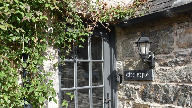 Um edifício de pedra com uma porta de madeira escura, uma placa com a inscrição "the BYRE" e um candeeiro fixado na parede.