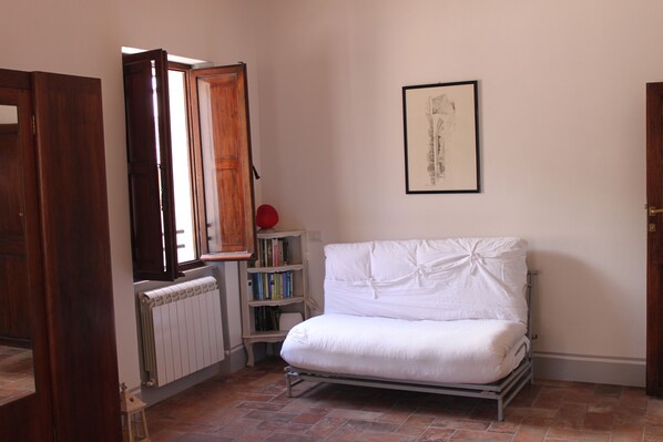 A room with a white mattress on a metal frame, a radiator, a bookshelf with books, and a framed picture on the wall.