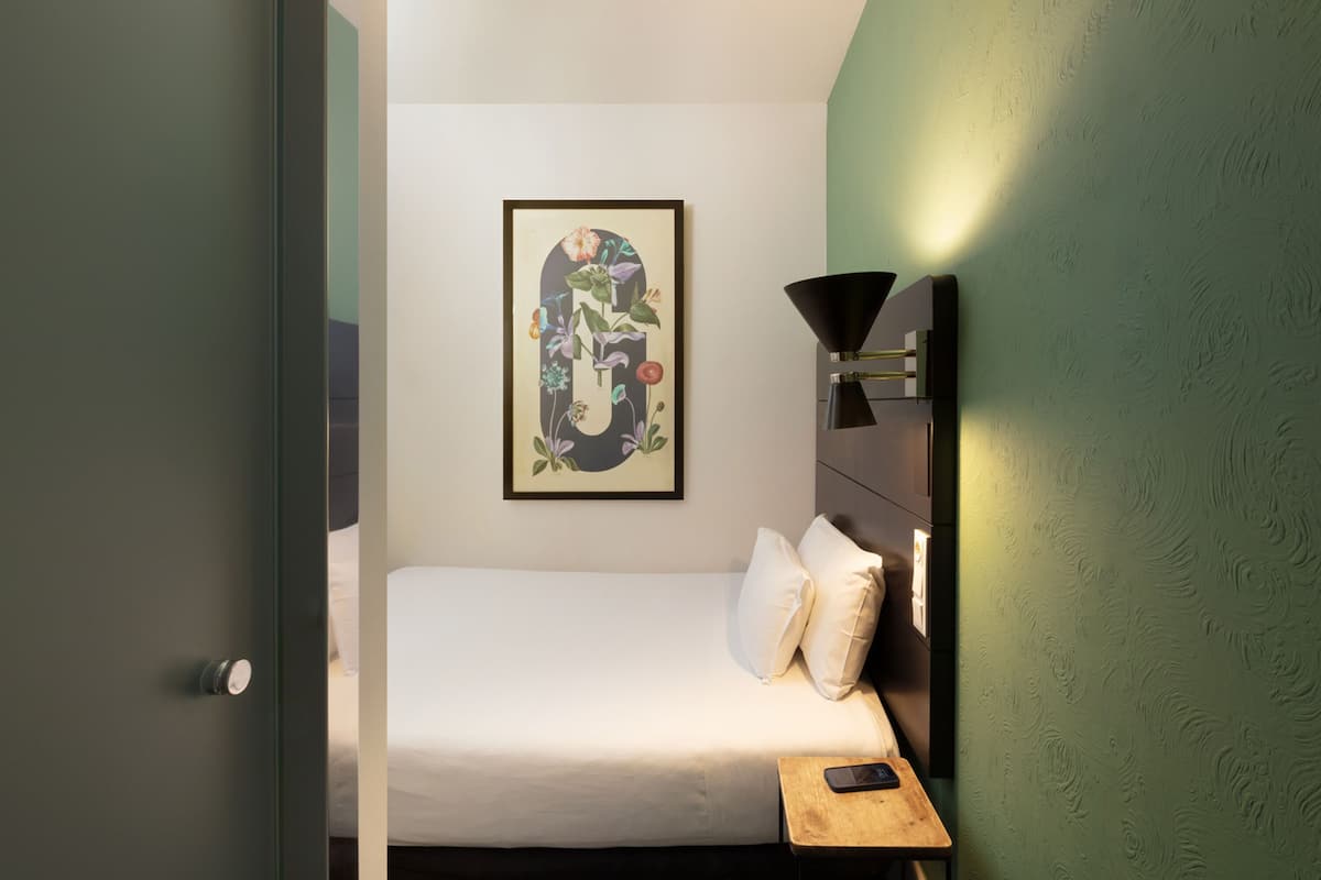 Une chambre d’hôtel bien rangée, avec un lit, une table de chevet sur laquelle se trouve un téléphone, une œuvre d’art florale encadrée accrochée au mur et une lampe fixée au mur.