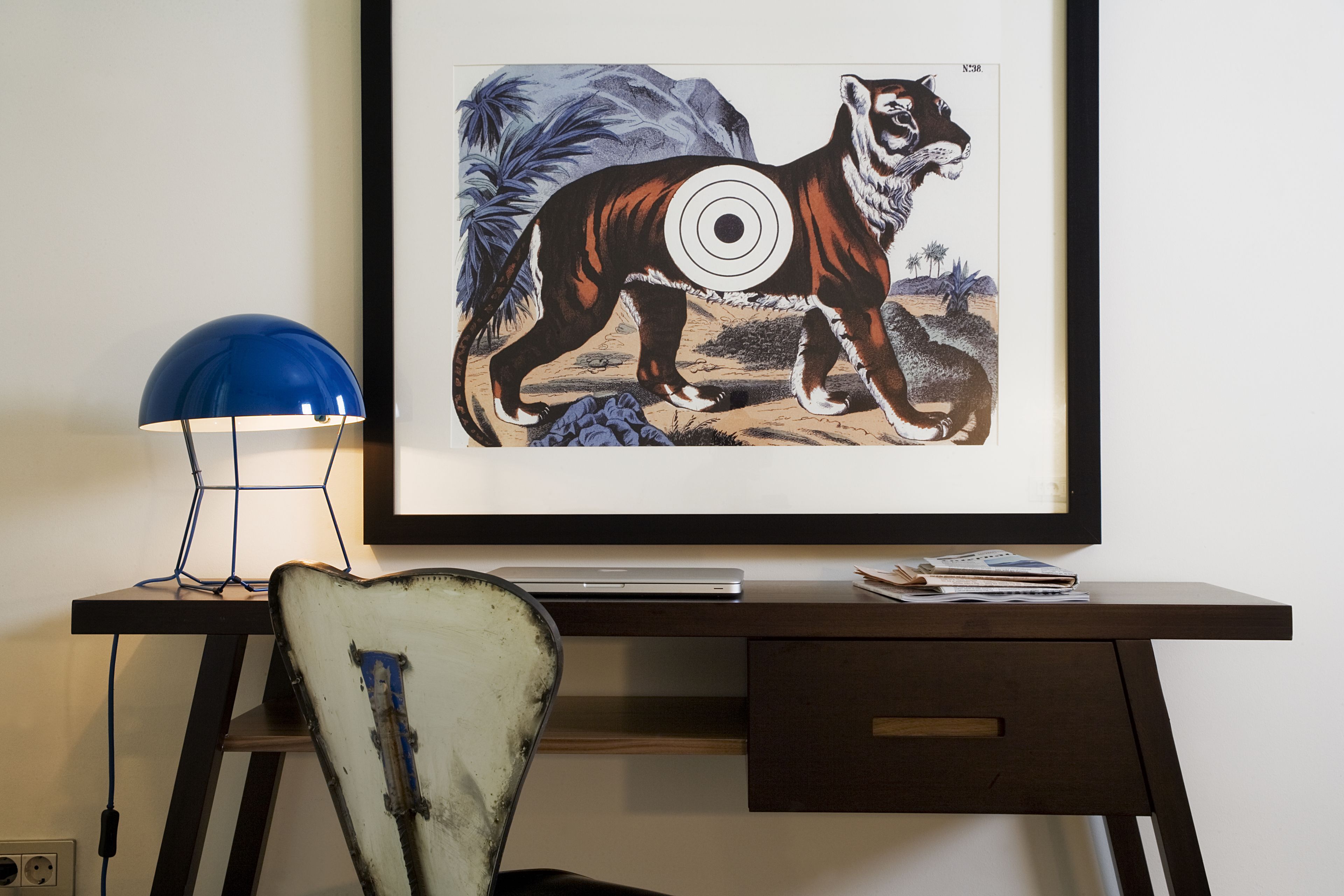 Un escritorio con una computadora portátil, una ilustración enmarcada de vida silvestre y una lámpara azul.