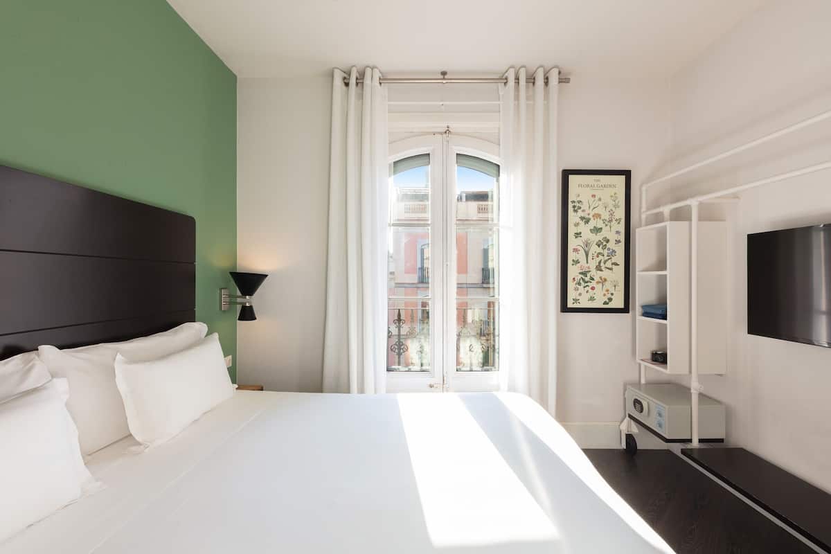 Une chambre d’hôtel avec un grand lit, une vue sur les bâtiments par la fenêtre, un téléviseur fixé au mur et une étagère avec divers objets.