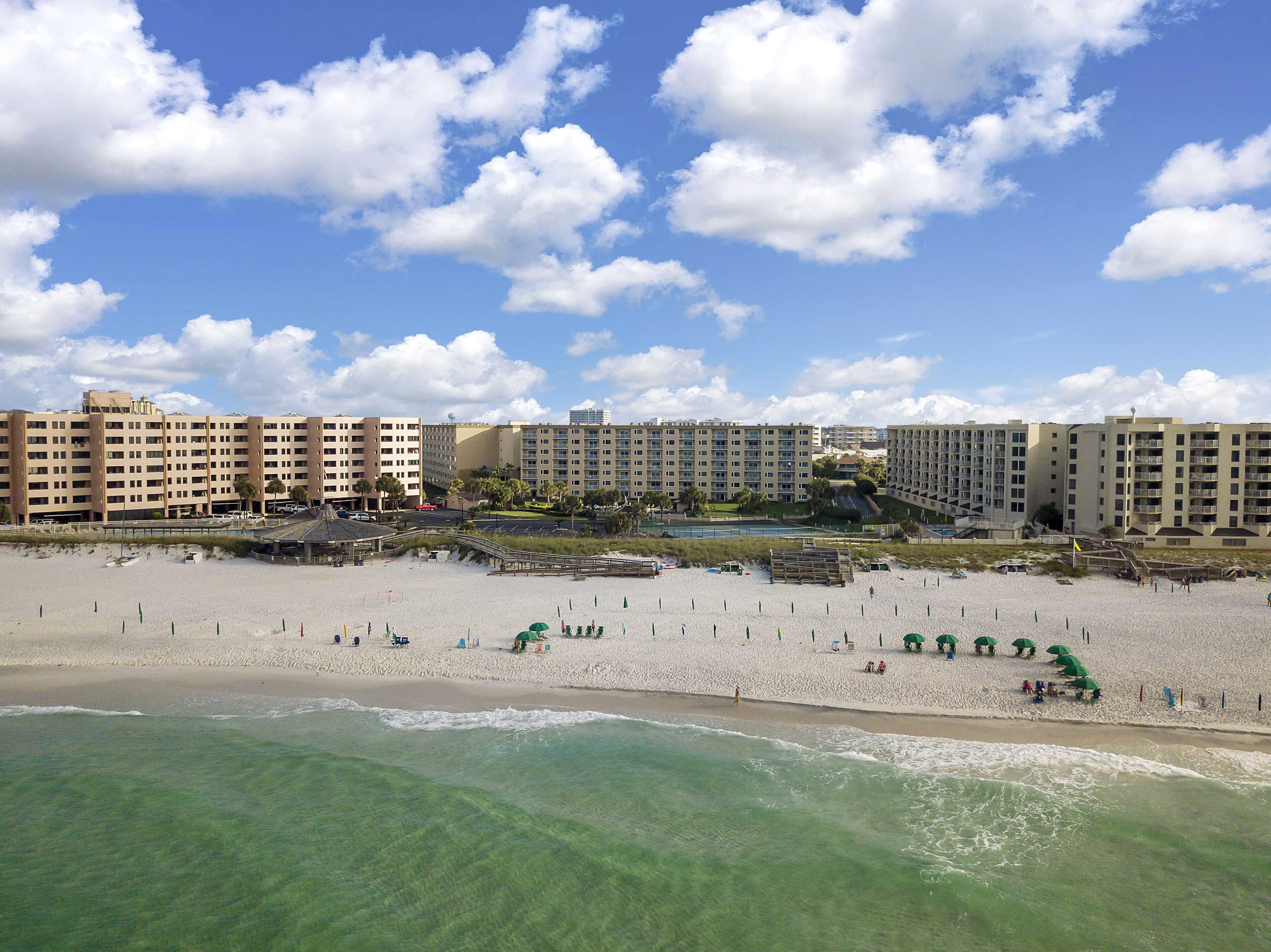 Un hôtel situé en bord de mer, composé de plusieurs bâtiments, avec une plage de sable fin et un ciel d’un bleu éclatant.