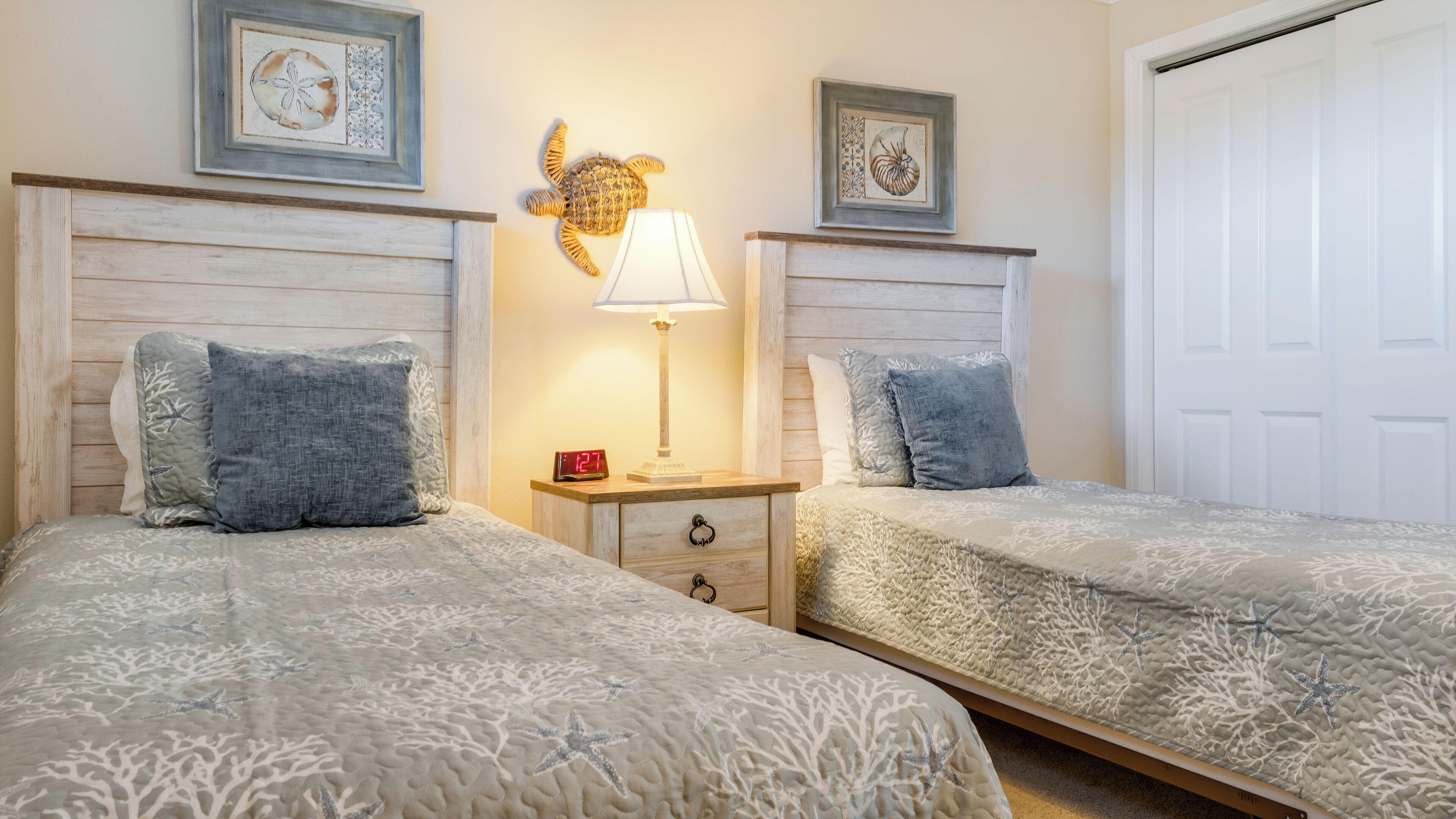 A bedroom with two beds, each with a headboard, a bedside table with a lamp, and framed artwork on the wall.