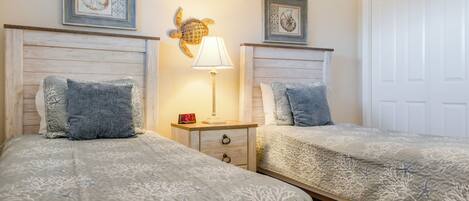 A bedroom with two beds, each with a headboard, a bedside table with a lamp, and framed artwork on the wall.